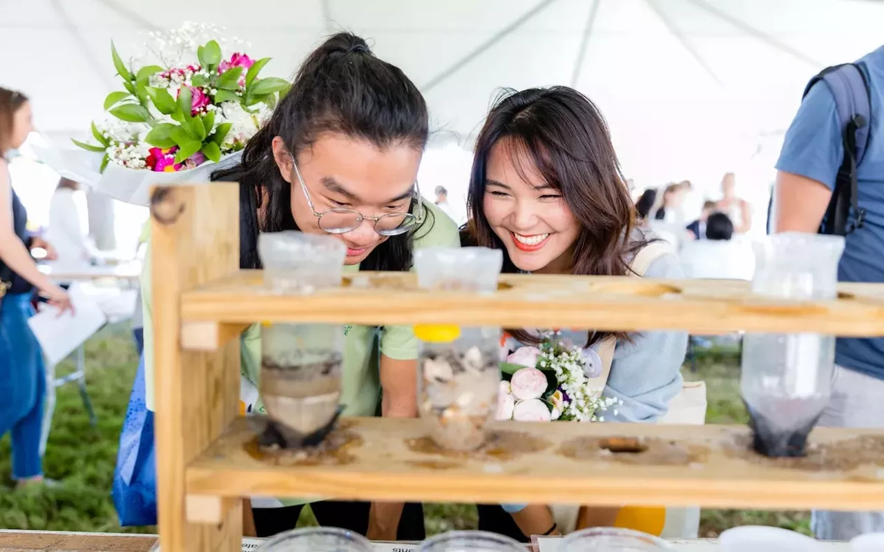 Two people, behind a test tube holder, holding tubes that contain seeds, dirt and plants.